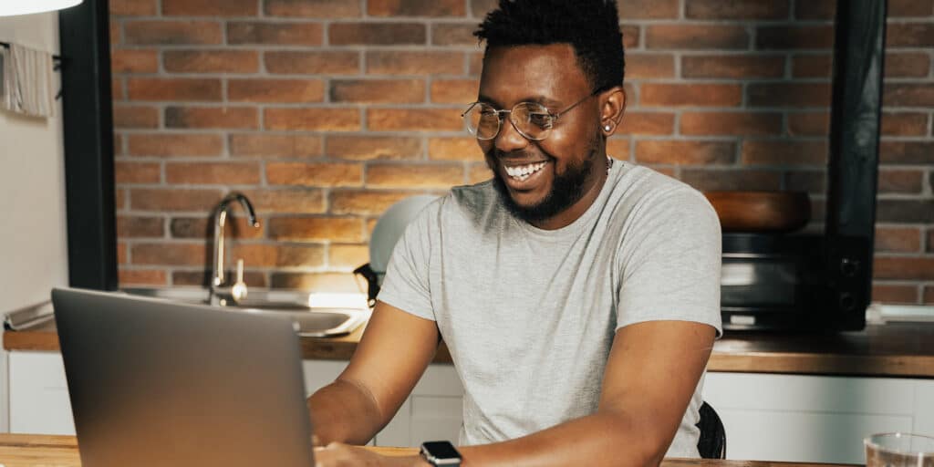 A smiling man sits at a workspace and types on an open laptop computer.