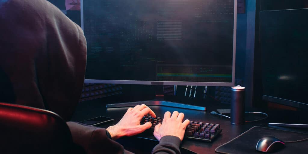 A criminal wearing a hooded sweatshirt is seated at a desktop computer typing on a keyword.