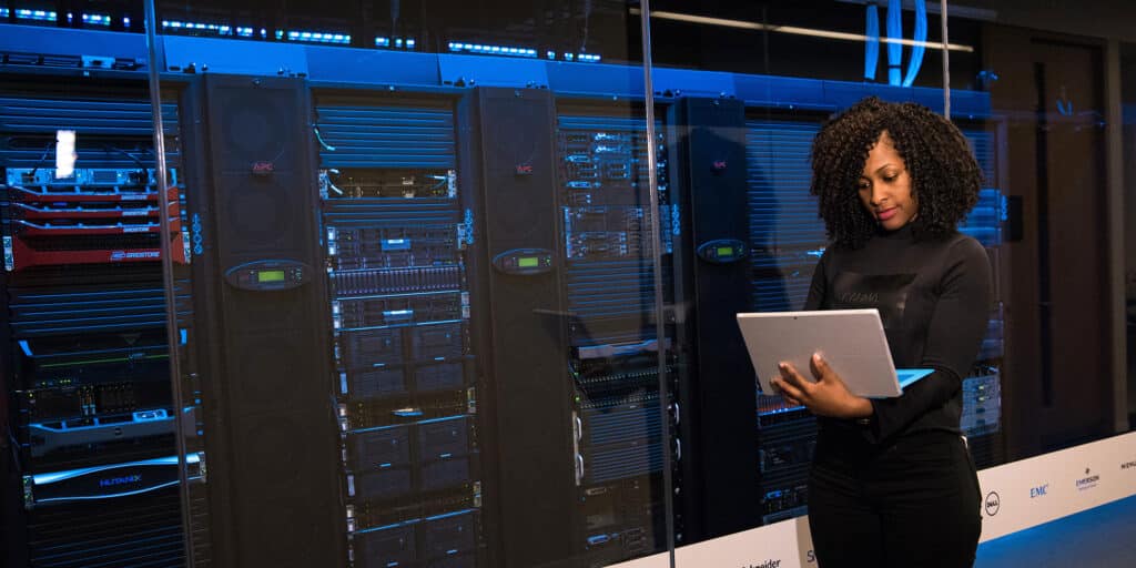 An IT worker in a data center standing next to a server rack and typing on a laptop computer.