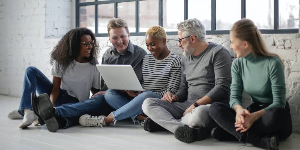 A team of five co-workers seated on the floor of an office look at content on a laptop.