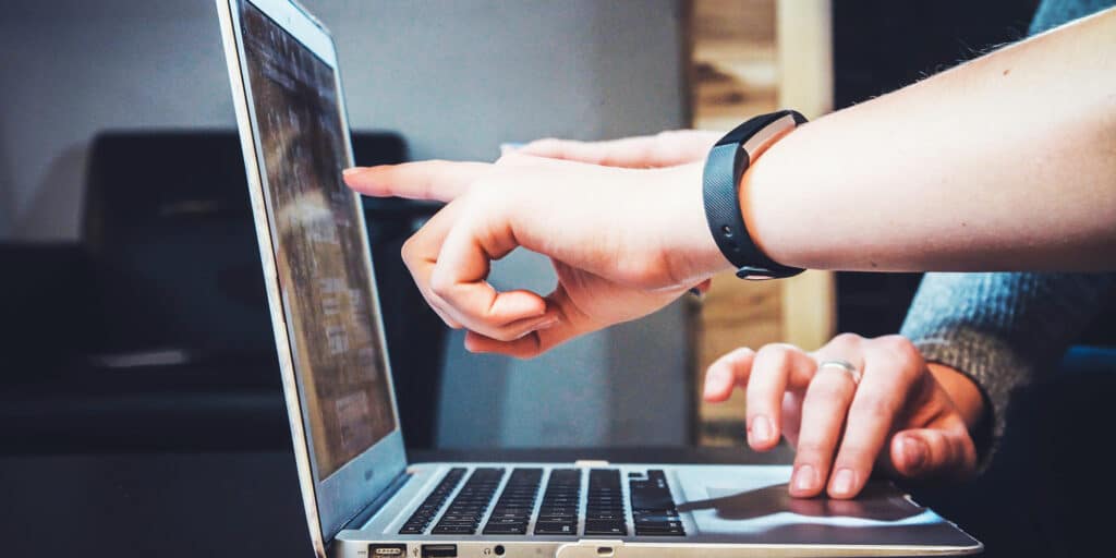 Close-up of an open laptop computer with one user's hand operating the trackpad and a different user's hand pointing at the screen.