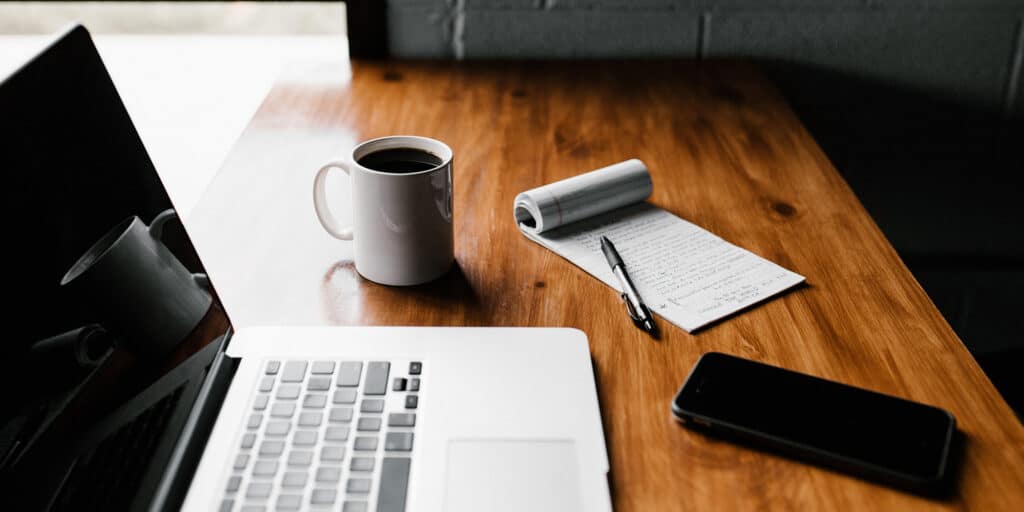 A full cup of coffee, a smartphone, and a pen and notepad on a desktop next to an open laptop computer.