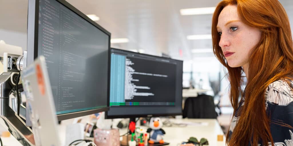 A woman sitting at a desk in front of an open laptop computer and two large displays.