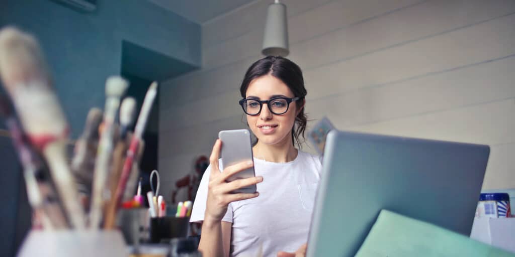 A woman wearing glasses seated at a laptop computer and looking at her smartphone.